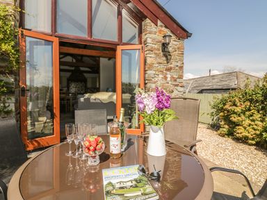 An outdoor patio with a round glass table holding a vase of flowers strawberries a bottle of drink glasses and a magazine in front of an open glass door at The Wagon House in Tresmeer near Launceston