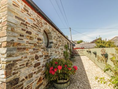 A stone wall with a round window beside a gravel path and flower pots attached to a wooden fence at The Wagon House in Tresmeer near Launceston