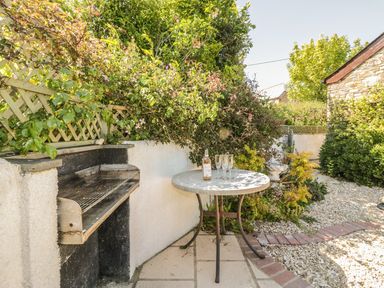 An outdoor patio area with a built in barbecue grill a round table with a bottle and glasses surrounded by plants at The Wagon House Tresmeer near Launceston
