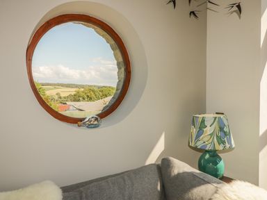 A round window with a model ship in a glass bottle on the sill above a gray sofa and a green table lamp with patterned shade at The Wagon House in Tresmeer near Launceston