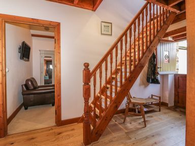 A wooden staircase in a hallway with a chair and a small shoe rack near the door and a leather armchair and wall-mounted television visible through an open doorway at The Wagon House in Tresmeer near Launceston