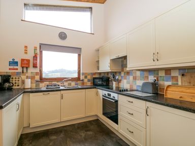 A kitchen with white cabinets black countertops a window with a blind a clock and various appliances at The Ash Lodge near Lyme Regis