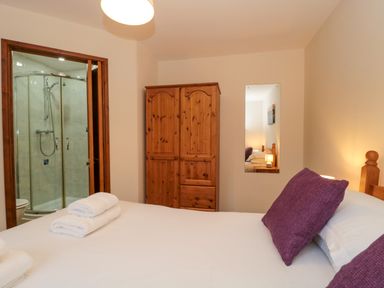 A bedroom with a bed, purple pillows, white towels on the bed, a wooden wardrobe, a mirror, and an en suite bathroom with a shower at The Ash Lodge near Lyme Regis