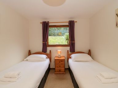 A bedroom with two single beds separated by a wooden nightstand with a lamp under a window with purple curtains at The Ash Lodge near Lyme Regis