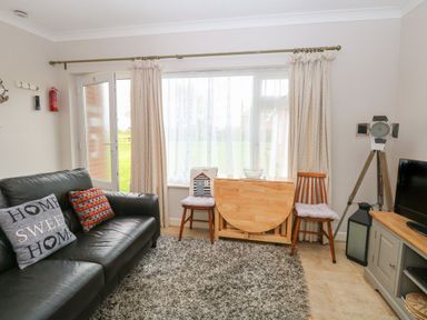A living room with a black leather sofa and cushions a wooden folding table two chairs a large window with curtains a gray carpet a tripod lamp and a television unit at En-Casa in Mundesley