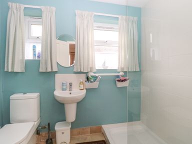 A bathroom with a toilet sink shower two windows with curtains and a small round mirror at En-Casa in Mundesley