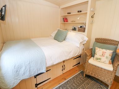 A small bedroom with a bed against the wall a wicker chair and built in shelves at Shepherds Hut in Castleton Peak District