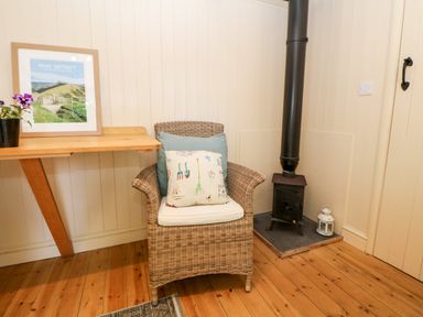 A wicker armchair with two cushions next to a wood stove and a wooden table with a framed peak district picture and potted plant at Shepherds Hut in Castleton Peak District