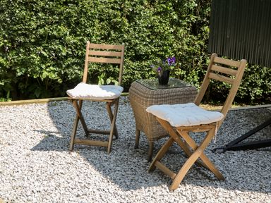 An outdoor seating area with two wooden chairs with cushions and a wicker table with a flower pot at Shepherd's Hut in Castleton Peak District