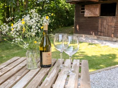 A wooden outdoor table with a bottle of wine flowers in a jar and two wine glasses near a wooden shed in a garden at Shepherd's Hut in Castleton Peak District