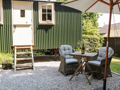 An outdoor seating area with two wicker chairs and a wooden table under an umbrella next to a green cabin called Shepherd's Hut in Castleton Peak District