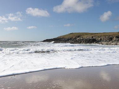 A sandy beach with waves and rocky cliffs under a blue sky at Plas Bach in Aberffraw