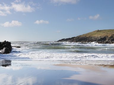 A beach with waves rolling in and a rocky hill under a blue sky at Plas Bach in Aberffraw