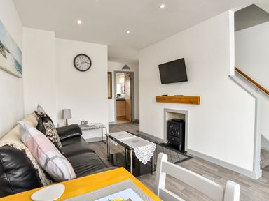A living room with a black leather sofa cushions a wall clock a wall mounted TV and a small stove at Plas Bach in Aberffraw
