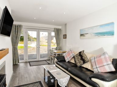 A living room with a black and white sofa with cushions a glass coffee table a wall-mounted television and double glass doors at Plas Bach in Aberffraw