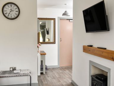 A hallway with a clock on the wall a bench a mirror a table with flowers a door a hanging light and a mounted television at Plas Bach in Aberffraw