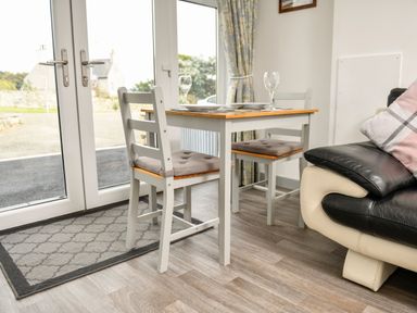A dining area with a wooden table and two chairs next to glass doors and a black and white sofa at Plas Bach in Aberffraw