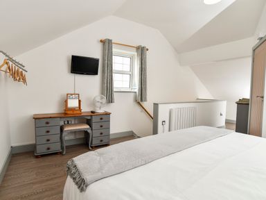 A bedroom with a bed, a grey and wood dressing table, a small stool, a wall-mounted TV, a window with curtains, and a rail with hangers at Plas Bach in Aberffraw