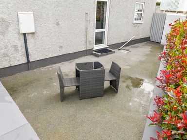 A patio area with a wicker table and four chairs near a door and a hedge at Plas Bach in Aberffraw