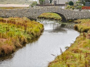 A stone arch bridge over a narrow river with grassy banks and houses in the background at Plas Bach in Aberffraw