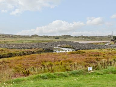 An arched stone bridge over a small stream with grass and bushes around at Plas Bach in Aberffraw