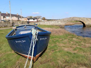 A blue boat named Beverley Jane on grass near a stone bridge over water with houses in the background at Plas Bach in Aberffraw