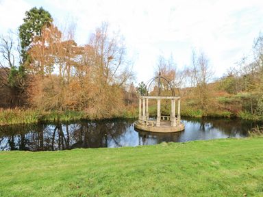 A pond with a circular stone gazebo in the middle surrounded by grass and trees at Snowdrop in Saltburn-by-the-Sea