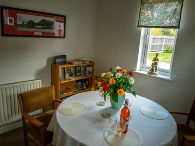 A dining room with a round table covered with a white tablecloth flowers in a vase on the table wooden chairs a window with floral blinds a small bookshelf with books and a figurine on the windowsill at Anvil Lodge in Goulceby near Louth