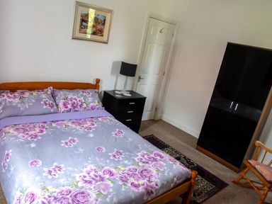 A bedroom with a wooden bed with floral bedding a black bedside table lamp a wooden rocking chair a dark wardrobe and a framed picture on the wall at Anvil Lodge in Goulceby near Louth