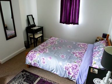A bedroom with a bed with floral bedding purple curtains a dressing table with a chair a wall mirror a bedside table with a lamp and a radio at Anvil Lodge in Goulceby near Louth