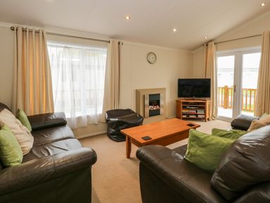 A living room with brown leather sofas a wooden coffee table a wall clock a tv on a wooden stand and cream curtains at Jolie in Hunstanton