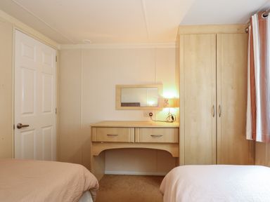 A bedroom with two beds a wooden dressing table with a mirror a lamp a wardrobe and a white door at Jolie in Hunstanton