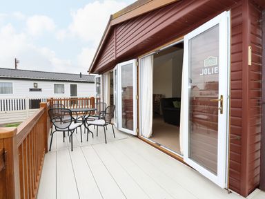 An outdoor deck with a metal table and four chairs next to an open door of a wooden cabin at Jolie in Hunstanton