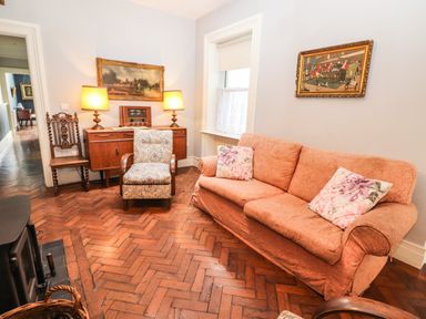 A living room with a peach sofa and floral cushions a patterned armchair a wooden sideboard with two lamps and a vintage radio two paintings on the wall and a wooden floor at The Merchant's Store in Kilrush Country Clare