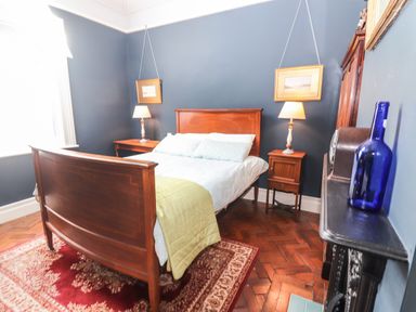 A bedroom with a wooden bed two bedside tables with lamps paintings on the wall a blue bottle on a mantelpiece and a red patterned rug at The Merchant's Store in Kilrush Country Clare