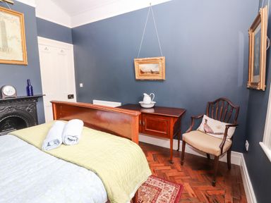 A bedroom with a wooden bed with folded towels on the bed a wooden side table with a pitcher and basin a wooden chair with a cushion and framed pictures on blue walls at The Merchant's Store in Kilrush Country Clare