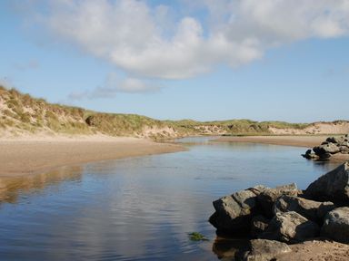 A sandy beach with a calm water stream and grass-covered dunes at Maelog 14 Tides Reach in Rhosneigr