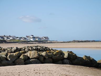 A rocky beach with large stones and water with houses in the background at Maelog 14 Tides Reach in Rhosneigr