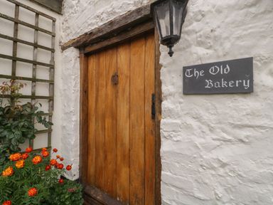 A wooden door with a black lantern and a sign that says The Old Bakery on a white textured wall with red and orange flowers nearby at The Old Bakery in Crowcombe