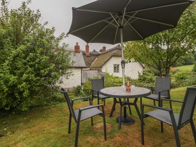 A garden with a round table set under a large umbrella with four chairs and a bottle with glasses on the table at The Old Bakery in Crowcombe