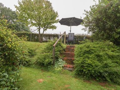 A garden with stairs leading to a patio table with chairs and an umbrella at The Old Bakery in Crowcombe