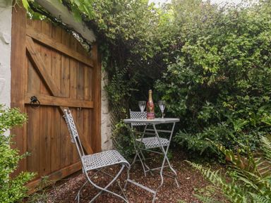A small outdoor seating area with a wooden gate metal table and two chairs surrounded by greenery at The Old Bakery in Crowcombe