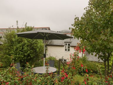 A garden with a table umbrella chairs flowers and a stone house in the background at The Old Bakery in Crowcombe