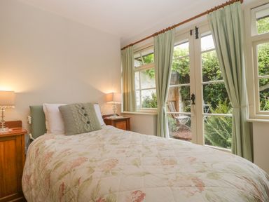 A bedroom with a bed covered in floral bedding two bedside tables with lamps and windows with curtains overlooking a garden at The Old Bakery in Crowcombe