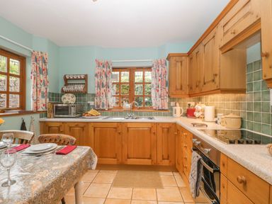A kitchen with wooden cabinets a dining table with plates and glasses and windows with patterned curtains at The Old Bakery in Crowcombe