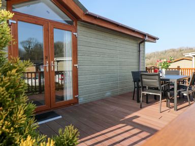 A wooden deck with a table and chairs outside a cabin with glass double doors at Tranquillity - Heritage Park Stepaside near Kilgetty