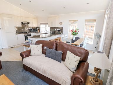 An open plan living area with a brown leather sofa, a dining table with chairs, and a kitchen with white cabinets at Tranquillity - Heritage Park in Stepaside near Kilgetty