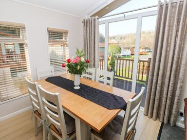 A dining area with a wooden table six white chairs a black table runner and a vase of flowers beside large sliding glass doors at Tranquillity - Heritage Park Stepaside near Kilgetty