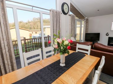 A dining table with a flower vase and white chairs near large windows in a living room at Tranquillity - Heritage Park Stepaside near Kilgetty