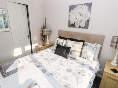 A bedroom with a bed with floral bedding cushions on the bed two bedside tables with lamps and a door on the left wall at Tranquillity - Heritage Park Stepaside near Kilgetty